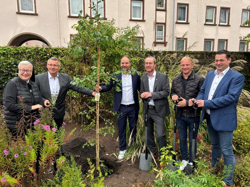 Fünf Männer und eine Nonne pflanzen gemeinsam einen jungen Baum in einem Garten. Sie stehen lächelnd mit Schaufeln in den Händen um das frisch eingepflanzte Bäumchen herum, umgeben von Pflanzen und Blumen. Im Hintergrund sind Wohngebäude zu sehen.