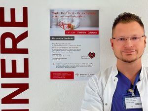 Ein Arzt in weißem Kittel und blauem Hemd steht lächelnd vor einer Wand mit der Aufschrift „INNERE“. Neben ihm hängt ein Plakat mit der Überschrift „Stärke Dein Herz – Herzschwäche erkennen und behandeln“. Der Arzt trägt eine Brille und hat ein Namensschild. Im Plakat wird eine Veranstaltung zur Herzgesundheit angekündigt, die am 13. November 2024 in der Cafeteria stattfindet.