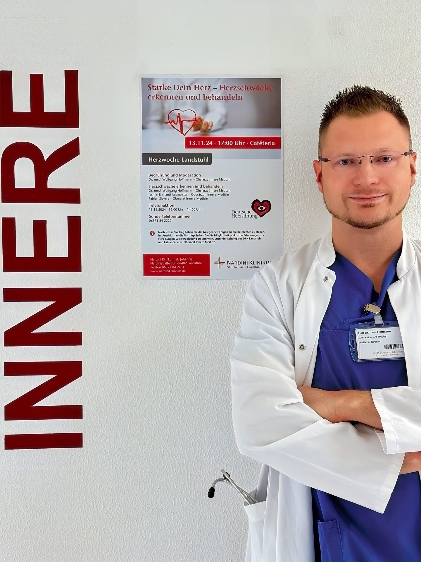 Ein Arzt in weißem Kittel und blauem Hemd steht lächelnd vor einer Wand mit der Aufschrift „INNERE“. Neben ihm hängt ein Plakat mit der Überschrift „Stärke Dein Herz – Herzschwäche erkennen und behandeln“. Der Arzt trägt eine Brille und hat ein Namensschild. Im Plakat wird eine Veranstaltung zur Herzgesundheit angekündigt, die am 13. November 2024 in der Cafeteria stattfindet.